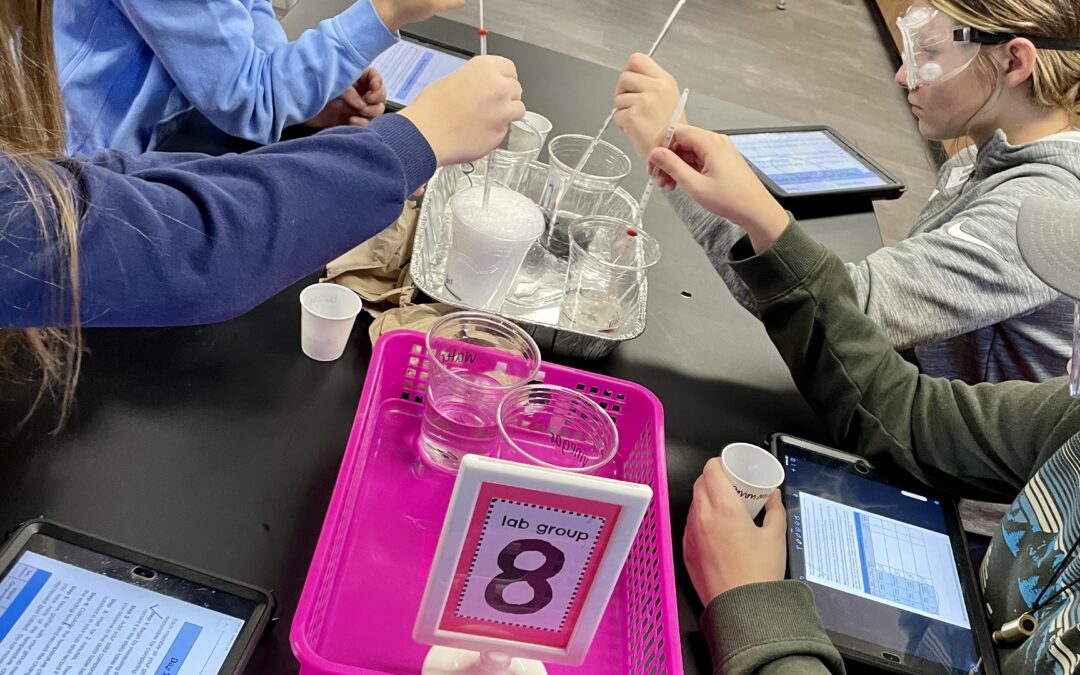 Four students seated at a lab table, wearing goggles, stirring mixtures in clear cups. A bright pink tray labeled "Lab Group 8" holds supplies in the center of the table.